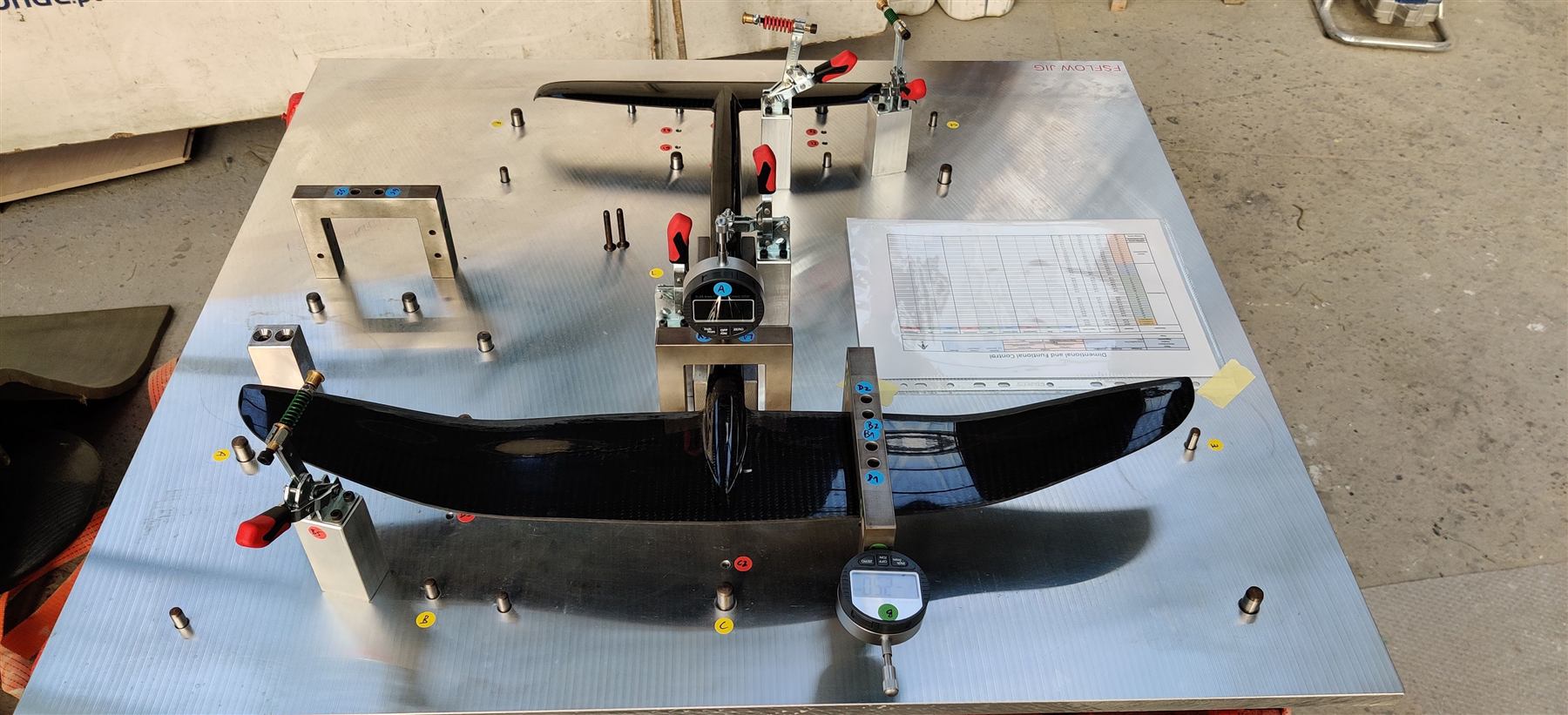 A precision inspection fixture on a granite surface plate, a carbon foil clamped in place, dial and digital gauges reading deviation at labelled datum points, dimensional-control checklist alongside.
