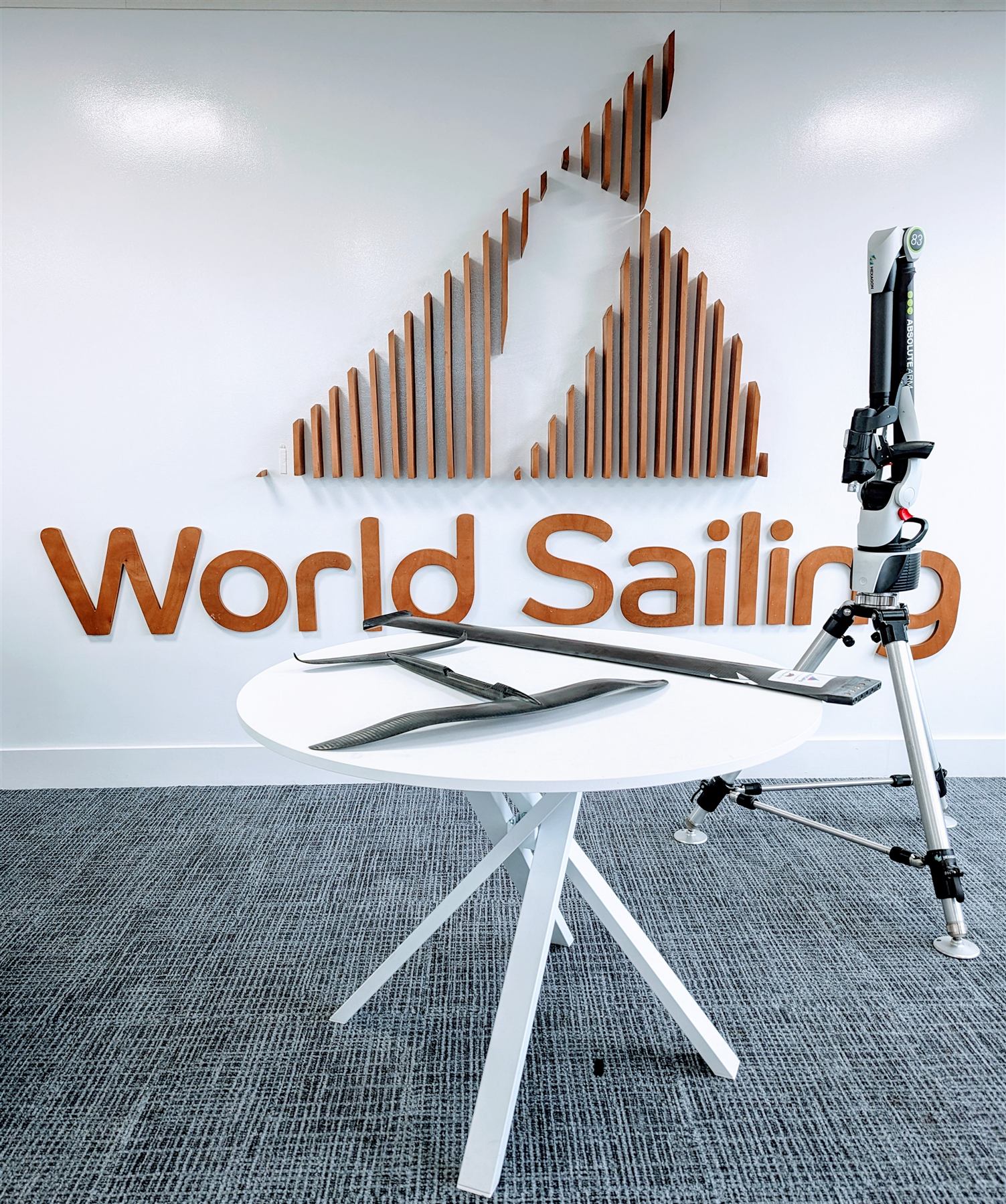 Two carbon hydrofoils on a table beneath the World Sailing logo, next to a portable CMM measuring arm, at World Sailing's London headquarters.
